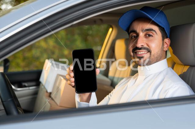 خدمات الشحن  الالكتروني الآمن, صورة مقربة لمندوب عربي خليجي سعودي مبتسم يرتدي الزي التقليدي و القبعة الزرقاء يجلس داخل السيارة و يقوم بعرض شاشة سوداء فارغة عبر الهاتف المحمول, استخدام التقنية الحديثة في توصيل الطلبات, النظر إلى الكاميرا بإيماءات الرضا و السرور