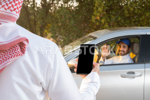 ايماءات القاء التحية والسلام، خدمات الديلفري السريعة في السعودية، صورة من الخلف لرجل عربي خليجي سعودي يمسك الهاتف المحمول بشاشة سوداء فارغة، الثقة المتبادلة بين العملاء وشركات الشحن، مفهوم التسوق الالكتروني، اداء العمل بجودة واتقان