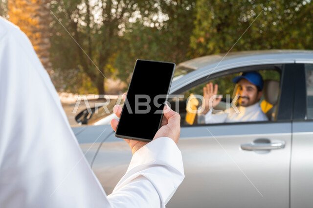 خدمات الديلفري السريعة في السعودية، صورة من الخلف لرجل عربي خليجي سعودي يمسك الهاتف المحمول بشاشة سوداء فارغة، القاء التحية والسلام، الثقة المتبادلة بين العملاء وشركات الشحن، مفهوم التسوق الالكتروني، اداء العمل بجودة واتقان