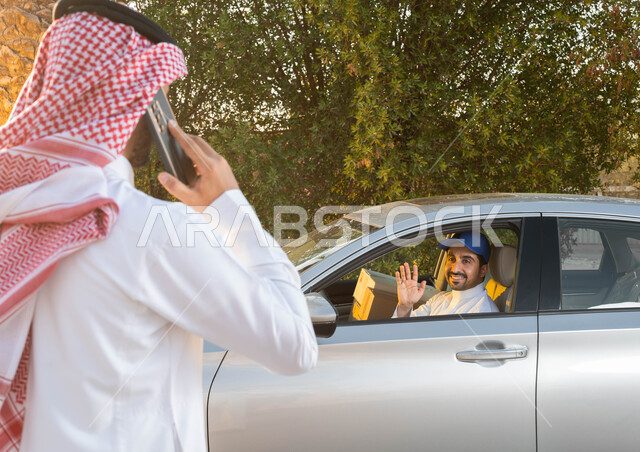 Providing greater comfort for individuals, the moment the delivery vehicle arrives, a picture from behind of a Saudi Gulf Arab man wearing traditional clothing holding a mobile phone, gestures of greeting and greetings, mutual trust between customers and shipping companies, fast and safe delivery service, performing work with quality and mastery.