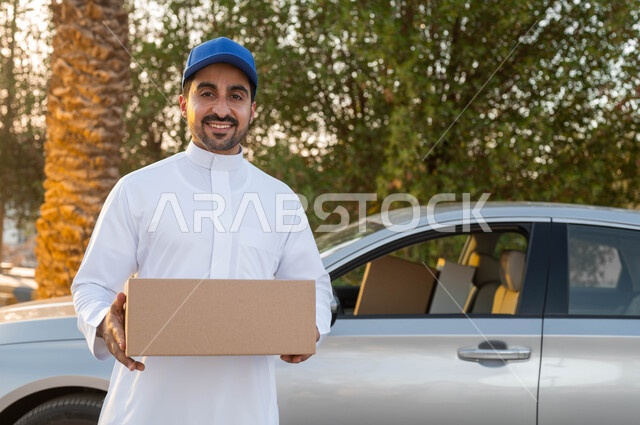 السرعة والتفاني في أداء العمل، مفهوم التسوق الالكتروني، خدمة الديلفري الآمنة، مندوب توصيل عربي خليجي سعودي يرتدي الزي التقليدي يقف أمام سيارة يقوم بحمل طرد لتسلميه، النظر الى الكاميرا بايماءات البهجة والسرور، تسليم البضائع والطلبات للزبائن