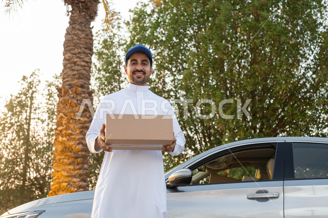 مفهوم التسوق الالكتروني، السرعة والتفاني في أداء العمل، خدمة الديلفري الآمنة، مندوب توصيل عربي خليجي سعودي يرتدي الزي التقليدي يقف أمام سيارة يقوم بحمل طرد لتسلميه، النظر الى الكاميرا بايماءات البهجة والسرور، تسليم البضائع والطلبات للزبائن
