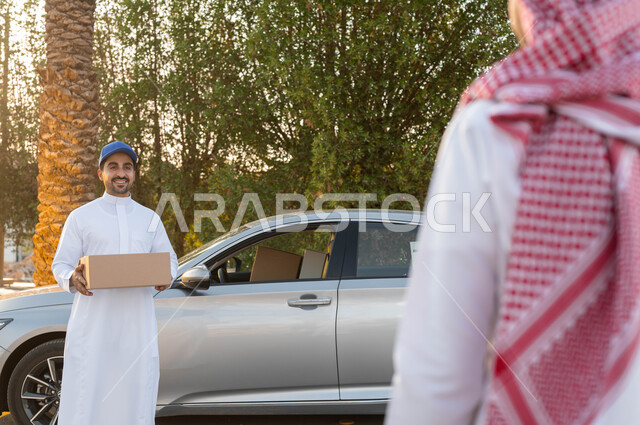 توصيل البضائع والطلبات للزبائن، خدمة الديلفري السريعة والآمنة، مندوب توصيل عربي خليجي سعودي يرتدي الزي التقليدي يقوم بحمل طرد لتسليمه ، الثقة المتبادلة بين العميل وشركات الشحن، مفهوم التسوق الالكتروني، الشراء عن طريق الانترنت