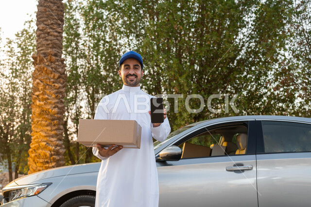 النظر الى الكاميرا بتعابير البهجة والسرور، خدمات الشحن والتوصيل السريعة الآمنة، عرض شاشة سوداء فارغة عبر الهاتف المحمول، مندوب توصيل عربي خليجي سعودي يقف أمام سيارة يحمل بيده الجوال و طرد التسليم، ميزة الشراء عن طريق الانترنت