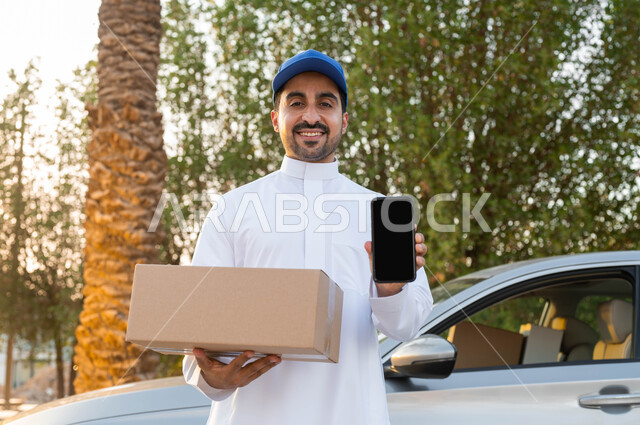 خدمات الشحن والتوصيل السريعة الآمنة، عرض شاشة سوداء فارغة عبر الهاتف المحمول، مندوب توصيل عربي خليجي سعودي يقف أمام سيارة يحمل بيده الجوال و طرد التسليم، مفهوم التسوق الالكتروني، النظر الى الكاميرا بتعابير البهجة والسرور