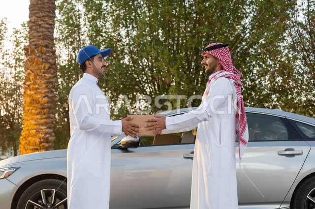 تعابير البهجة والسرور عند وصول الطلبيات، الشراء عن طريق الانترنت، مفهوم التسوق الالكتروني، خدمات التوصيل السريع، رجل عربي خليجي سعودي يرتدي الثوب التقليدي والشماغ يقوم باستلام طرد من مندوب الشحن، خدمة الديلفري السريعة والآمنة، تسليم البضائع والطلبات للزبائن