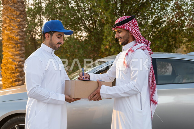 ايماءات الرضا والسرور، مفهوم التسوق الالكتروني، رجل عربي خليجي سعودي يرتدي الثوب التقليدي والشماغ يقوم بالتوقيع لاستلام طرد من مندوب الشحن، خدمة الديلفري السريعة والآمنة، الشراء عن طريق الانترنت، تسليم البضائع والطلبات للزبائن