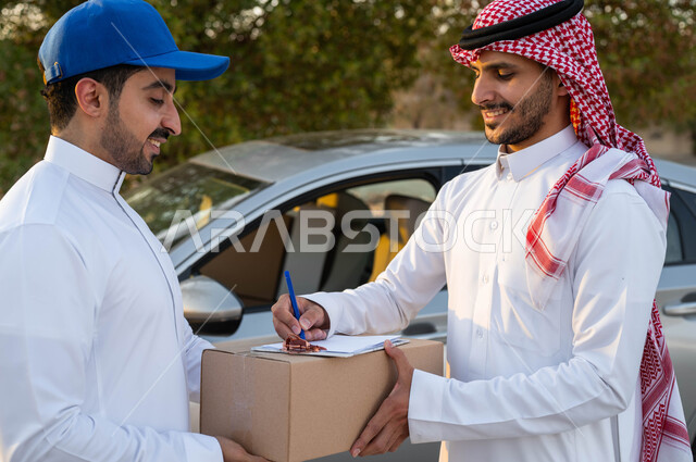 تسليم البضائع والطلبات للزبائن، ايماءات الرضا والسرور، مفهوم التسوق الالكتروني، صورة مقربة لرجل عربي خليجي سعودي يرتدي الثوب التقليدي والشماغ يقوم بالتوقيع لاستلام طرد من مندوب الشحن، خدمة الديلفري السريعة والآمنة، الشراء عن طريق الانترنت