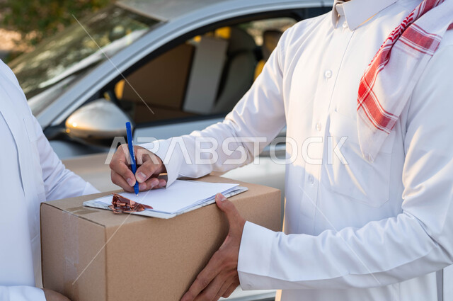 خدمات التوصيل السريع، صورة مقربة لأيدي رجل عربي خليجي سعودي يرتدي الثوب التقليدي والشماغ يقوم بالتوقيع لاستلام طرد من مندوب الشحن، خدمة الديلفري السريعة والآمنة، الشراء عن طريق الانترنت، حمل صناديق المشتريات، مفهوم التسوق الالكتروني