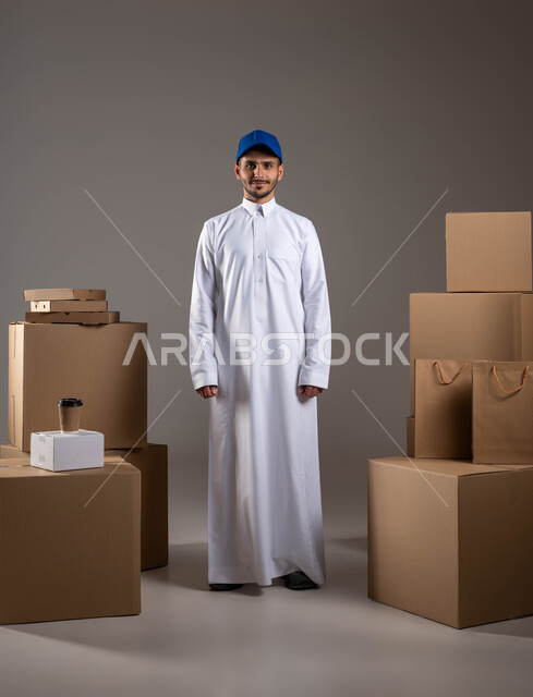 مفهوم الآمان في نقل الطلبيات، توفير راحة أكبر للافراد من خلال الطلب اونلاين، النظر إلى الكاميرا بإيماءات الرضا والسرور، خدمات الشحن ذات الجودة العالية، بورتريه لمندوب توصيل عربي خليجي سعودي يرتدي الزي التقليدي يقف باستقامة بتعابير الثقة بالنفس، خلفية رمادية