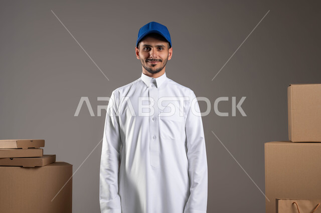 Providing greater comfort for individuals by ordering online, looking at the camera with gestures of satisfaction and pleasure, high-quality shipping services, the concept of safety in transporting orders, a portrait of a Saudi Arabian Gulf delivery representative wearing traditional clothing, standing straight with an expression of self-confidence, gray background.