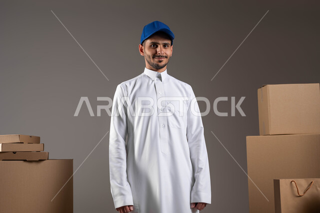 النظر إلى الكاميرا بإيماءات الرضا والسرور، خدمات الشحن ذات الجودة العالية، مفهوم الآمان في نقل الطلبيات، توفير راحة أكبر للافراد من خلال الطلب اونلاين، بورتريه لمندوب توصيل عربي خليجي سعودي يرتدي الزي التقليدي يقف باستقامة بتعابير الثقة بالنفس، خلفية رمادية