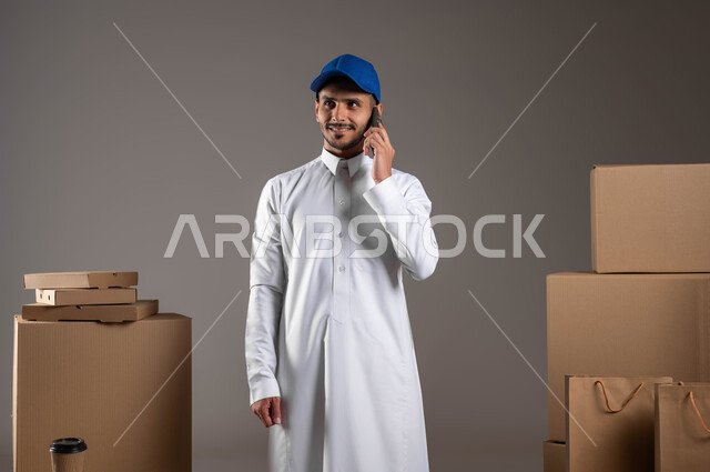 التواصل الفعال مع العملاء، مفهوم الآمان في نقل طلبيات الاونلاين، بورتريه مقرب لشاب عربي خليجي سعودي يرتدي الزي التقليدي والقبعة الزرقاء يقوم باجراء مكالمة هاتفية عبر الهاتف المحمول، ايماءات الرضا والسرور، خدمة التوصيل السريع في السعودية، خلفيه رمادية