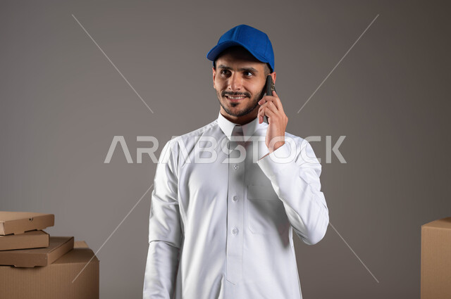 مفهوم الآمان في نقل طلبيات الاونلاين، بورتريه مقرب لشاب عربي خليجي سعودي يرتدي الزي التقليدي والقبعة الزرقاء يقوم باجراء مكالمة هاتفية عبر الهاتف المحمول، التواصل الفعال مع العملاء، ايماءات الرضا والسرور، خدمة التوصيل السريع في السعودية، خلفيه رمادية