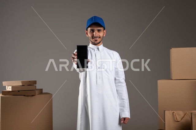 عرض شاشة سوداء فارغة عبر الهاتف المحمول، النظر إلى الكاميرا بايماءات الرضا والسرور، بورتريه لمندوب توصيل عربي خليجي سعودي يرتدي الزي التقليدي والقبعة الزرقاء، استخدام التكنولوجيا الحديثة في الحياة العملية، الشراء عن طريق الانترنت، خلفيه رمادية