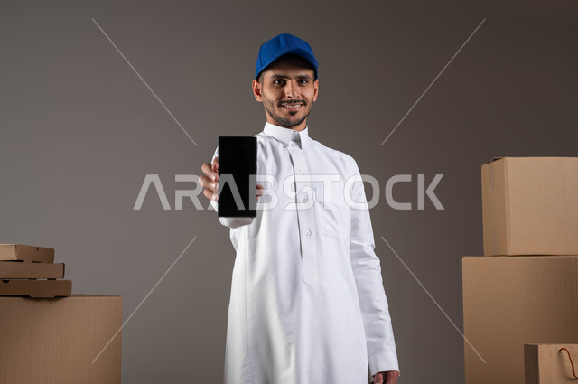 النظر إلى الكاميرا بايماءات الرضا والسرور، بورتريه لمندوب توصيل عربي خليجي سعودي يرتدي الزي التقليدي والقبعة الزرقاء، عرض شاشة سوداء فارغة عبر الهاتف المحمول، استخدام التكنولوجيا الحديثة في الحياة العملية، الشراء عن طريق الانترنت، خلفيه رمادية