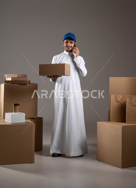 التواصل مع العملاء، استخدام التكنولوجيا الحديثة في الحياة العملية، الشراء عن طريق الانترنت، بورتريه لمندوب توصيل عربي خليجي سعودي يرتدي الزي التقليدي والقبعة الزرقاء يحمل بيده صندوق وينظر الى الكاميرا، اجراء مكالمة هاتفية عبر الهاتف المحمول، خلفيه رمادية