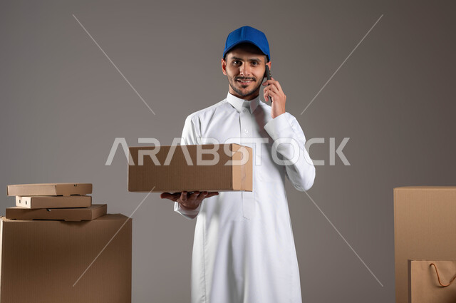 استخدام التكنولوجيا الحديثة في الحياة العملية، الشراء عن طريق الانترنت، بورتريه لمندوب توصيل عربي خليجي سعودي يرتدي الزي التقليدي والقبعة الزرقاء يحمل بيده صندوق وينظر الى الكاميرا، التواصل مع العملاء، اجراء مكالمة هاتفية عبر الهاتف المحمول، خلفيه رمادية
