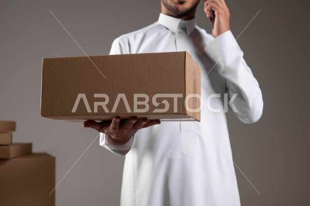 تسليم المشتريات والطلبات للزبائن، الاتصال بالعميل لاخباره بوصول طلبه, بورتريه لرجل عربي خليجي سعودي يرتدي الثوب التقليدي يقوم بأجراء مكالمه على الهاتف، خدمة ديلفري آمنة وموثوقة لضمان رضا العملاء، خلفيه رماديه