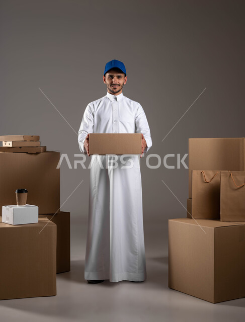 Providing a safe delivery service to ensure that orders reach customers safely, a portrait of a Saudi Gulf Arab man wearing a traditional dress and a blue hat, high-quality wrapped cartons to ensure the safety of shipments during transportation, a full-length body portrait, gray background.