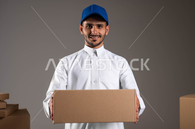 توصيل الطلبيات في الوقت المحدد وبشكل سليم، بورتريه مقرب لشاب عربي خليجي سعودي يرتدي الثوب التقليدي وقبعه زرقاء يمسك بيده كرتونه مغلقه، النظر الى الكاميرا بثقه وسعادة لوصول الطلبيه بسرعة، خلفيه رماديه
