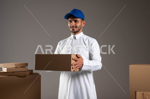 الكراتين المغلفة لتسهيل عمليات التخزين والتوزيع، بورتريه لرجل عربي سعودي خليجي مبتسم يرتدي الزي التقليدي وقبعه زرقاء يحمل بيده علبه كرتونيه، توفير خدمة التوصيل الآمنة لضمان وصول الطلبيات إلى العملاء بشكل سليم، خلفيه رماديه
