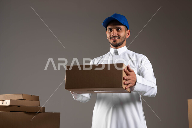 الكراتين المغلفة لحماية البضائع خلال عمليات الشحن والتخزين، بورتريه مقرب لرجل عربي سعودي خليجي مبتسم يرتدي الزي التقليدي وقبعه زرقاء يعمل في توصيل الطلبات والبضائع للزبائن,  خدمة ديلفري آمنة وموثوقة لضمان رضا العملاء، خلفيه رماديه