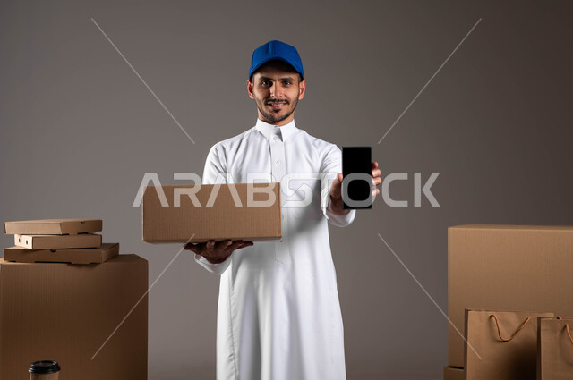 ايماءت الاعجاب باستخدام التقنيه والتكنولوجيا الحديثه، بورتريه لرجل عربي خليجي سعودي مبتسم يرتدي الثوب التقليدي وقبعة زرقاء يحمل بيده جوال بشاشة سوداء فارغة، كراتين وأكياس ورقية جاهزة للتوصيل، خلفيه رماديه