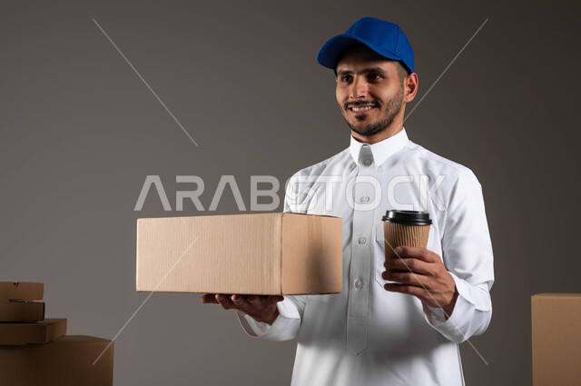 إيماءات الاستمتاع بتناول المشروبات الساخنة، بورتريه مقرب لشاب عربي خليجي سعودي يرتدي الزي التقليدي وقبعه زرقاء يحمل بيده كوب ورقي، كراتين الشحن والتوصيل الاونلاين، خلفيه رماديه