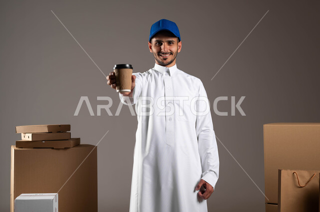 إيماءات الاستمتاع بتناول المشروبات الساخنة، بورتريه لشاب عربي سعودي خليجي يرتدي الثوب التقليدي وقبعه زرقاء يحمل بيده كوب ورقي، شراء القهوة من المقاهي السعوديه، صناديق كرتون وأكياس ورقية، خلفيه رماديه
