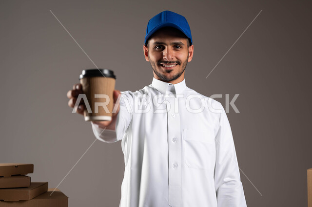 خدمات التوصيل السريع والامن للمشروبات الساخنة, شراء القهوة من المقاهي السعوديه، بورتريه مقرب لرجل عربي سعودي خليجي يرتدي الثوب التقليدي وقبعه زرقاء يحمل بيده كوب ورقي، النظر الى الكاميرا بثقه وسرور، خلفيه رماديه