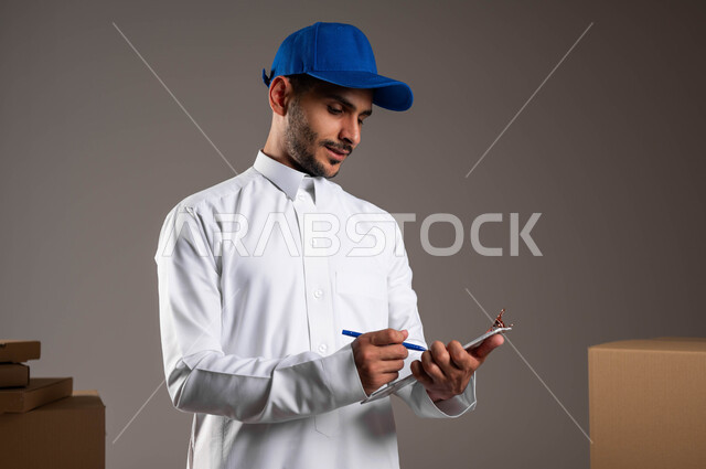 تسجيل طلبات العملاء بدقة لضمان وصولها كاملة وبشكل سليم، بورتريه مقرب لرجل عربي خليجي سعودي مبتسم يرتدي الثوب التقليدي وقبعه زرقاء يقوم بكتابة المنتجات المطلوبة على الورقة، خدمات شحن وتوصيل موثوقة وفعالة، خلفيه رماديه
