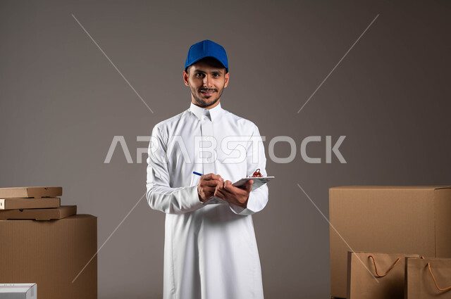 ايماءات الثقه والسرور لوصول الطلبيه بالوقت المحدد، بورتريه لرجل عربي خليجي سعودي مبتسم يرتدي الثوب التقليدي وقبعه زرقاء يقوم بالكتابة على الأوراق، تسجيل الطلبات بدقة من قبل عامل التوصيل، خلفيه رماديه
