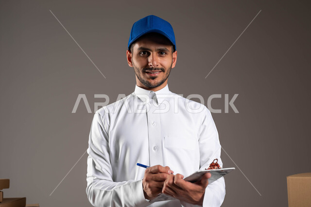 Adherence to the written timetable to ensure that the order arrives on time. A close-up portrait of a smiling Saudi Arabian Gulf man wearing traditional clothes and wearing a blue hat. He writes on paper. Orders are recorded accurately by the delivery worker, gray background.