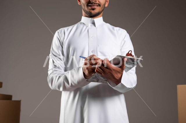 تسجيل وتدوين الطلبات بدقة من قبل عامل التوصيل، بورتريه لرجل عربي خليجي سعودي يرتدي الثوب التقليدي يقوم بالكتابة على الورق، الالتزام بالجدول الزمني المكتوب لضمان وصول الطلبية بالوقت المحدد، تسليم المشتريات للزبائن، خلفيه رماديه