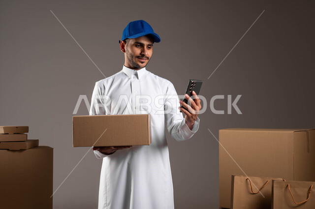 دمج العمل مع التقنية والتكنولوجيا, شركات التوصيل الاونلاين في المملكة, متابعة حالة الطلبات عبر تطبيقات الهاتف المحمول، بورتريه لشاب عربي خليجي سعودي يرتدي الثوب التقليدي وقبعه زرقاء يمسك بيده صندوق ويستخدم الجوال، خلفيه رماديه