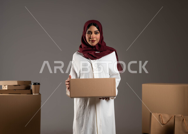 ايماءات السعادة  لوصول الطلبيه بشكل سليم، بورتريه لامراه عربية خليجية سعودية مبتسمه ترتدي عباءه بيضاء وحجاب أحمر تمسك بيدها صندوق، كراتين واكياس ورقيه جاهزة للشحن، جودة شركات التوصيل في المملكة، خلفيه رماديه