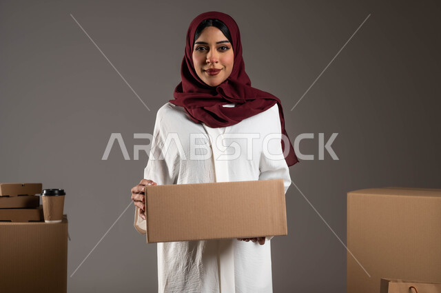Fast and safe delivery services, close-up portrait of a smiling Saudi Arabian Gulf woman wearing a white abaya and red hijab, holding a box in her hand, wrapped cartons in a variety of sizes and shapes, gestures of happiness that the order arrived safely, gray background