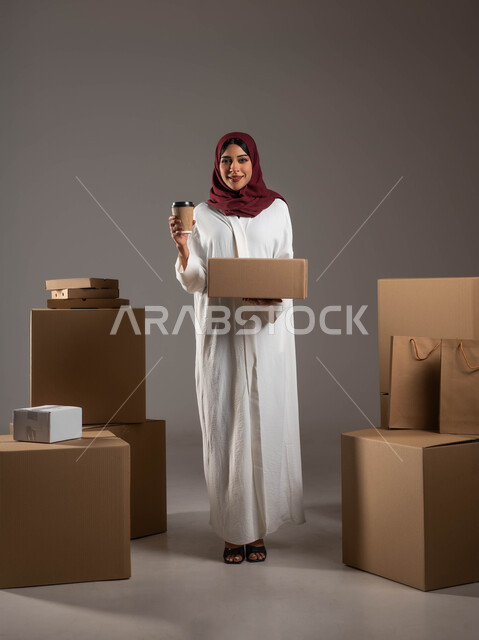 الكراتين المغلفة لحماية البضائع خلال عمليات الشحن والتخزين، بورتريه لامراه عربية خليجية سعودية مبتسمه ترتدي عباءه بيضاء وحجاب احمر تمسك بيدها كوب من المشروب الساخن، صوره لكامل الجسم، خلفيه رمادية