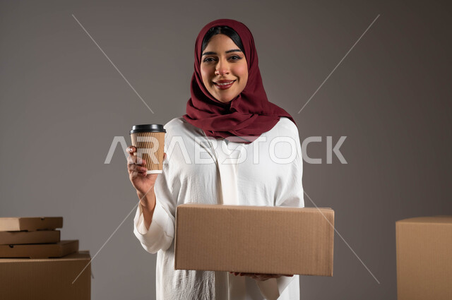 الكراتين المغلقة لتنظيم وترتيب المواد والمنتجات بشكل فعال، بورتريه مقرب لامراه عربية خليجية سعودية مبتسمه ترتدي عباءه بيضاء وحجاب أحمر تحمل بيدها صندوق، شركات توصيل المشروبات الساخنة، خلفيه رماديه