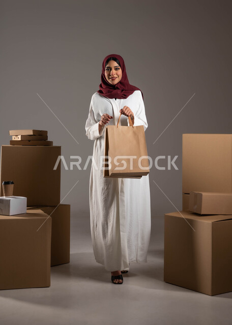 خدمات توصيل المنتجات السريعة والموثوقة، صدق وجودة شركات الشحن في المملكة، عملية نقل الطلبات للزبائن، بورتريه لامرأة عرية خليجية سعوية ترتدي عباءة بيضاء وحجاب خمري تحمل بيدها كيس كرتوني، صناديق كرتون التعبئة والتغليف، الطول الكامل للجسم، خلفية رمادية