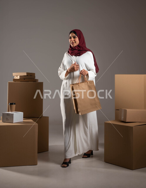 صدق وجودة شركات الشحن في المملكة، عملية نقل الطلبات للزبائن، خدمات توصيل المنتجات السريعة والموثوقة، بورتريه لامرأة عرية خليجية سعوية ترتدي عباءة بيضاء وحجاب خمري تحمل بيدها كيس كرتوني، صناديق كرتون التعبئة والتغليف، الطول الكامل للجسم، خلفية رمادية
