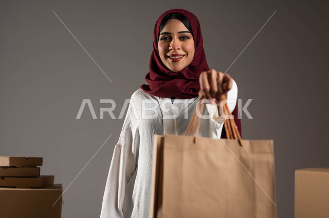 جودة وصدق شركات الشحن في المملكة، عملية نقل الطلبات للزبائن، خدمات توصيل المنتجات السريعة والموثوقة، بورتريه مقرب لامرأة عرية خليجية سعوية ترتدي عباءة بيضاء وحجاب خمري تحمل بيدها كيس كرتوني، صناديق كرتون التعبئة والتغليف، خلفية رمادية