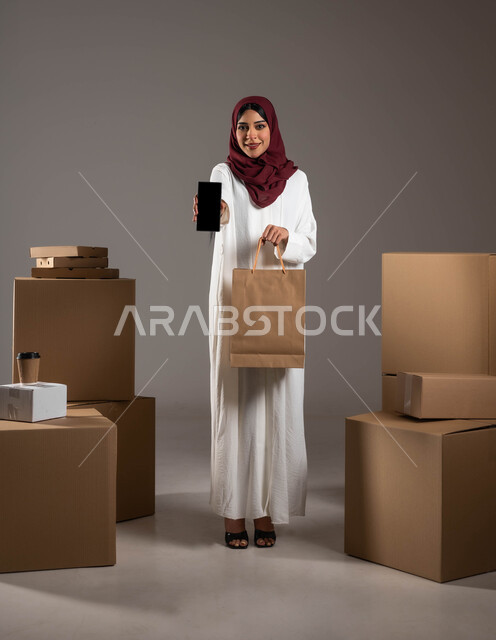 الشراء والتسوق الأونلاين، أداء العمل بدقة وإتقان، طلب المنتجات عبر تطبيقات الهاتف المحمول، بورتريه لامرأة عربية خليجية سعودية ترتدي عباءة بيضاء وحجاب أحمر، عرض شاشة سوداء فارغة عبر الموبايل، خدمات الشحن ذات الجودة العالية، الطول الكامل للجسم، خلفية رمادية