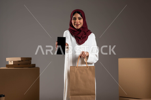 التسوق والشراء الأونلاين، أداء العمل بدقة وإتقان، طلب المنتجات عبر تطبيقات الهاتف المحمول، بورتريه لامرأة عربية خليجية سعودية ترتدي عباءة بيضاء وحجاب أحمر، عرض شاشة سوداء فارغة عبر الموبايل، خدمات الشحن ذات الجودة العالية، خلفية رمادية