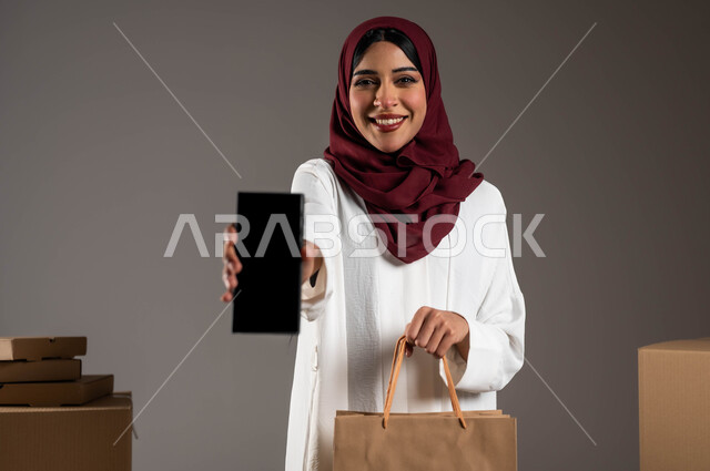 خدمات الشحن ذات الجودة العالية، التسوق والشراء الأونلاين، أداء العمل بدقة وإتقان، طلب المنتجات عبر تطبيقات الهاتف المحمول، بورتريه مقرب لامرأة عربية خليجية سعودية ترتدي عباءة بيضاء وحجاب أحمر، عرض شاشة سوداء فارغة عبر الموبايل، خلفية رمادية