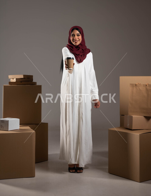 التسوق الذكي عبر الانترنت، النظر الى الكاميرا بايماءات السعادة والرضا، خدمات التوصيل السريع لضمان وصول المشروبات الساخنة، بورتريه لامرأة عربية خليجية سعودية ترتدي عباءة بيضاء وحجاب أحمر تقوم بحمل كأس القهوة، الطول الكامل للجسم، خلفية رمادية