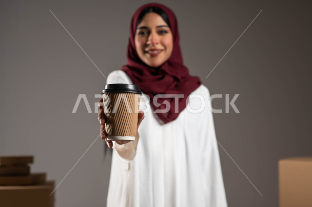 التسويق والترويج للمنتجات، خدمات التوصيل السريع لضمان وصول المشروبات الساخنة، بورتريه مقرب لامرأة عربية خليجية سعودية ترتدي عباءة بيضاء وحجاب أحمر تقوم بحمل كأس القهوة، التسوق الذكي عبر الانترنت، النظر الى الكاميرا بايماءات السعادة والرضا، خلفية رمادية