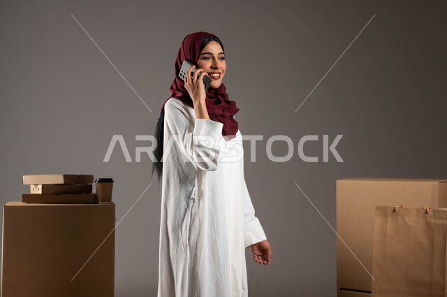 التواصل الفعال مع العملاء، إجراء المكالمات الهاتفية لتأكيد الحجوزات، بورتريه لامرأة عربية خليجية سعودية ترتدي عباءة بيضاء وحجاب أحمر تعمل في شركة توصيل، التسوق الذكي وطلب المنتجات أونلاين، خدمات الشحن السريعة والموثوقة، خلفية رمادية