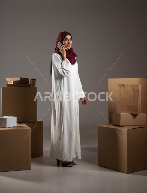 التسوق الذكي وطلب المنتجات أونلاين، إجراء المكالمات الهاتفية لتأكيد الحجوزات، التواصل الفعال مع العملاء، بورتريه لامرأة عربية خليجية سعودية ترتدي عباءة بيضاء وحجاب أحمر تعمل في شركة توصيل، خدمات الشحن السريعة والموثوقة، الطول الكامل للجسم، خلفية رمادية